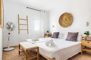 Ein Hotelzimmer mit zwei Betten, einem Standventilator, einer Holzleiter, einem gewebten Wandbehang und einem kleinen Tisch mit einer Pflanze.