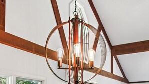 Una habitación con techo de madera, una lámpara colgante con bombillas, dos paneles decorativos en la pared, un sofá, una mesa con sillas y una ventana con vistas a follaje.