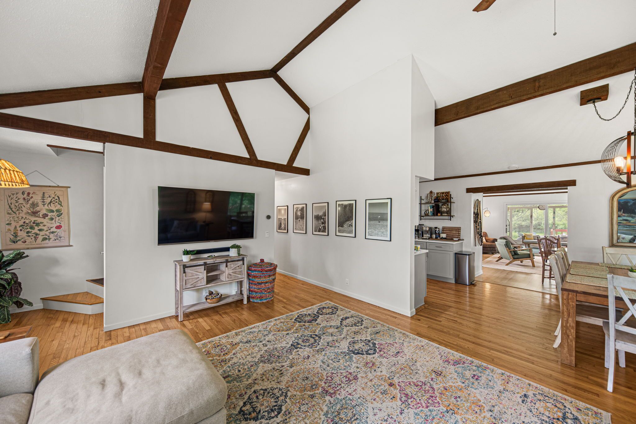 A spacious living room with a large flat-screen TV, wooden beams, a patterned rug, and a dining area with a wooden table and chairs.