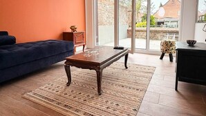 A living room with a dark blue sofa, a wooden coffee table, a patterned rug, and a view through large glass doors to the outside.