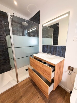 A modern bathroom with a glass shower enclosure, a white sink with wooden cabinet drawers, and a large mirror.