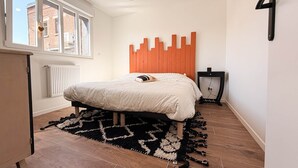 A bedroom with a bed, a radiator, a window, a wall-mounted number, and a hanging light fixture.