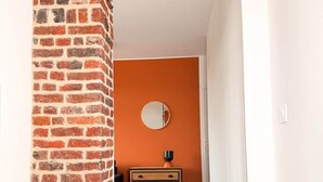 Ein Flur mit einer Ziegelssäule, einer orangefarbenen Wand, einem runden Spiegel und einer Holzkommode.