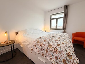 A bedroom with a bed, a nightstand, a lamp, a window, and an orange chair.
