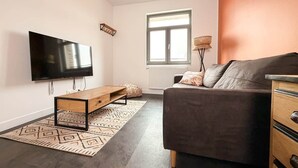 Ein modernes Wohnzimmer mit Flachbildfernseher, einer Couch, einem Couchtisch aus Holz, einem gemusterten Teppich und einem Fenster mit Jalousien.