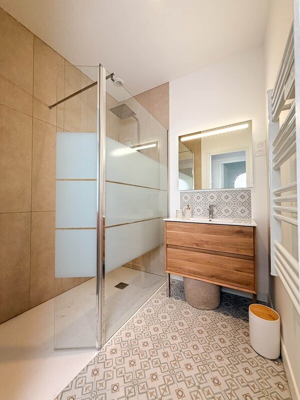 Un baño moderno con cabina de ducha de vidrio, tocador de madera, piso estampado y espejo con iluminación integrada.