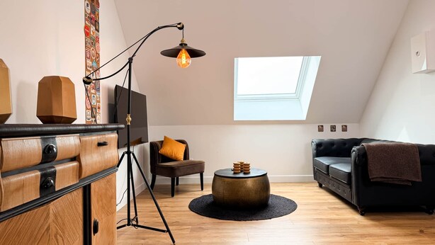 Um quarto em estilo loft com teto alto, claraboia, armário de madeira, sofá de couro preto, mesa de centro redonda e luminária de chão.