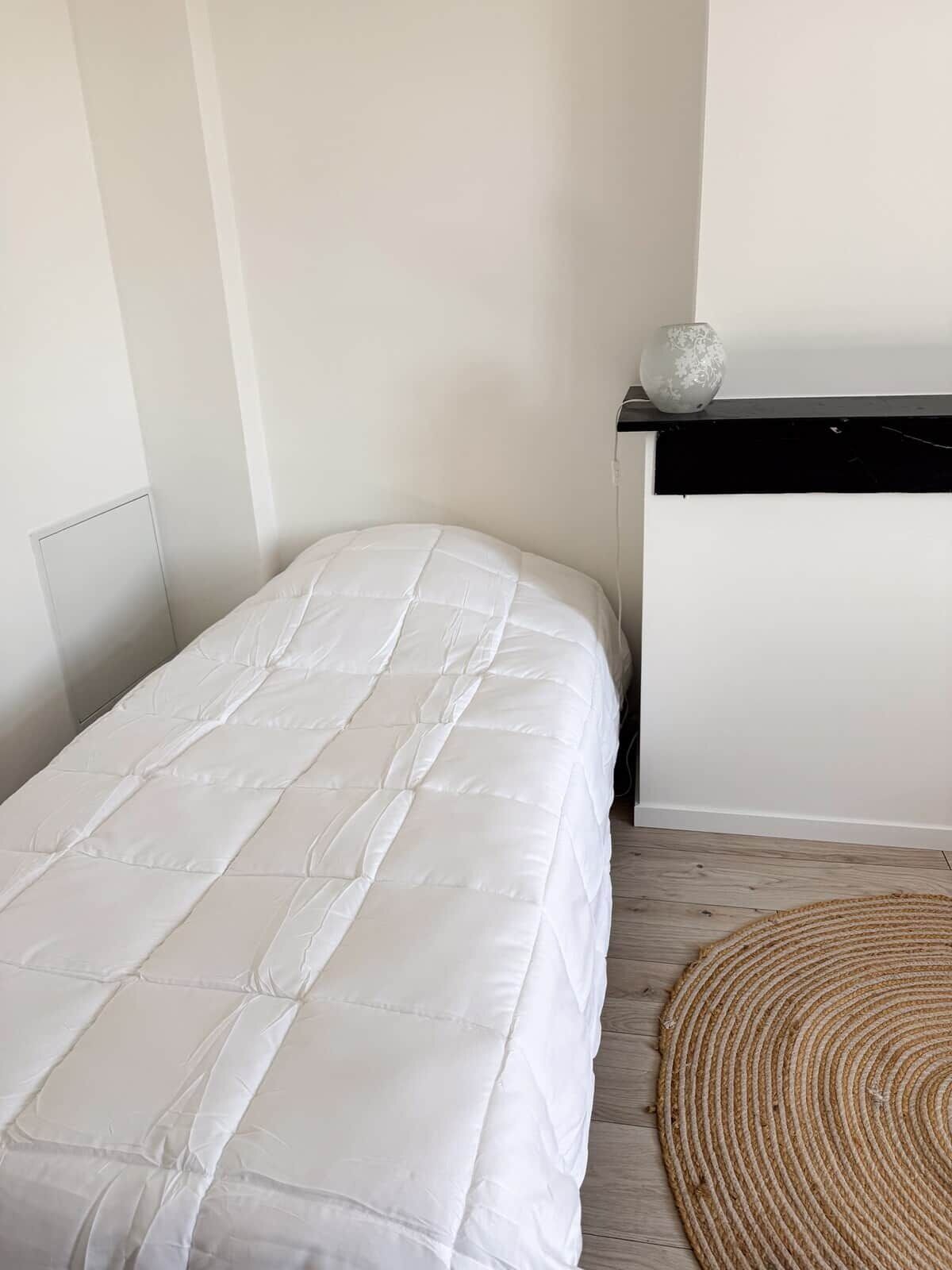 A bed with a white quilt, a small round rug, and a wall-mounted shelf with a decorative item.