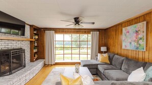 Un salon avec une cheminée, un ventilateur de plafond, une grande fenêtre avec des rideaux, un canapé gris et un tableau au mur.