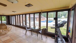 Une terrasse couverte équipée d'un tapis de course, de fauteuils en osier, d'une table en verre et de grandes fenêtres donnant sur un jardin avec une piscine.