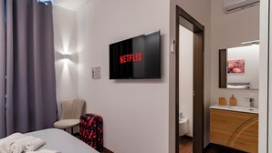 Habitación de hotel con cama, silla, maleta, televisor mostrando 'NETFLIX', baño con espejo y lavabo, y aire acondicionado instalado en la pared.
