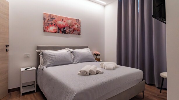 Una cama bien tendida con almohadas, una mesita de noche con control remoto y una obra de arte colgada en la pared.