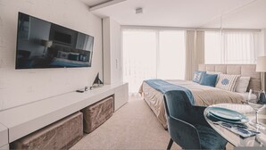 A modern hotel room with a large flat-screen TV mounted on a white brick wall, a bed with blue and beige bedding, a bedside table with a lamp, and a window with sheer curtains. - Marea Widok na Morze Studio Premium Hotel Seaside (gmina Parzęczew)