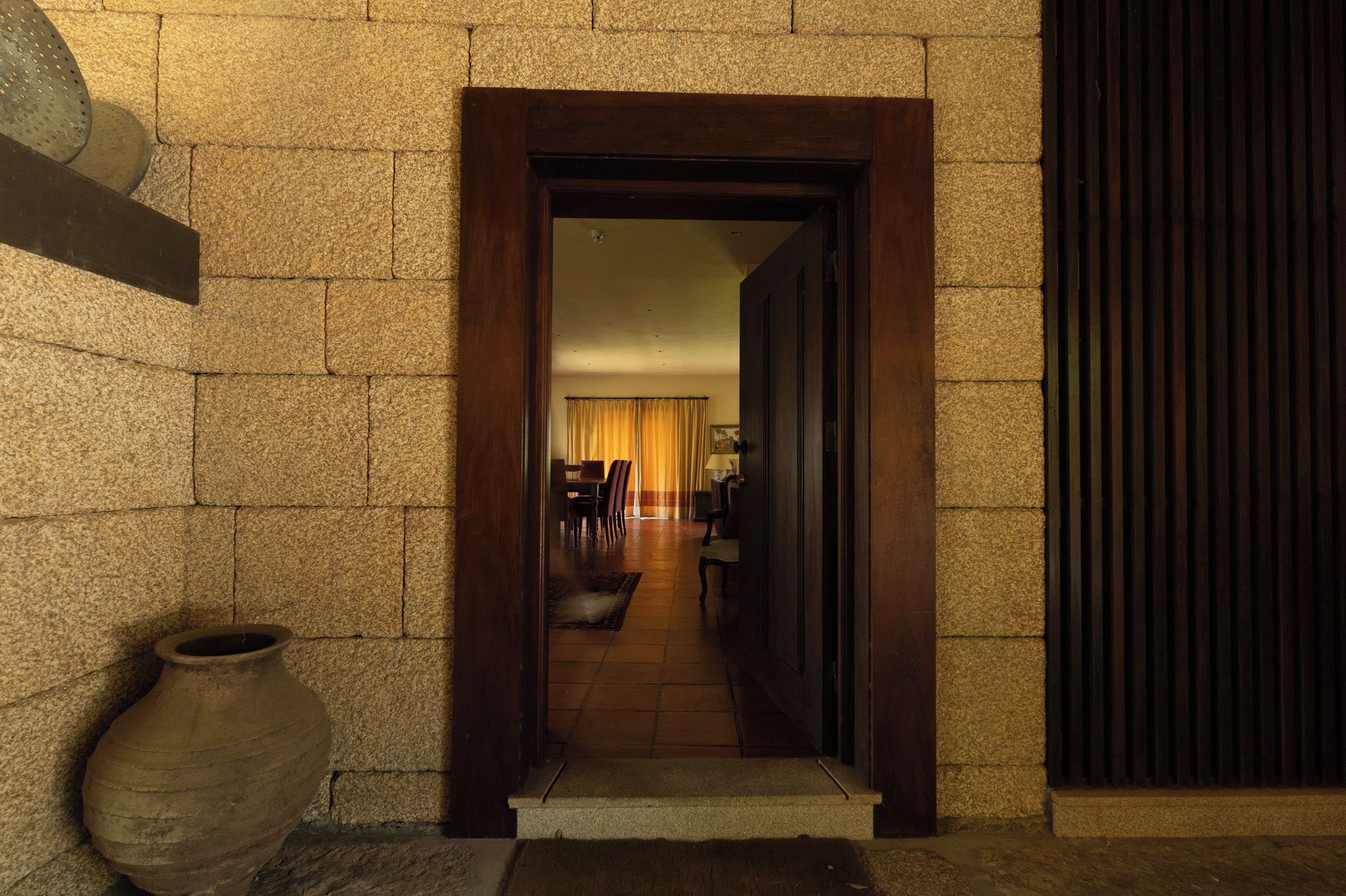 A hallway leading to a room with a wooden door, stone walls, and a large vase on the floor.