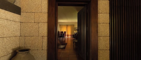 A hallway leading to a room with a wooden door, stone walls, and a large vase on the floor.