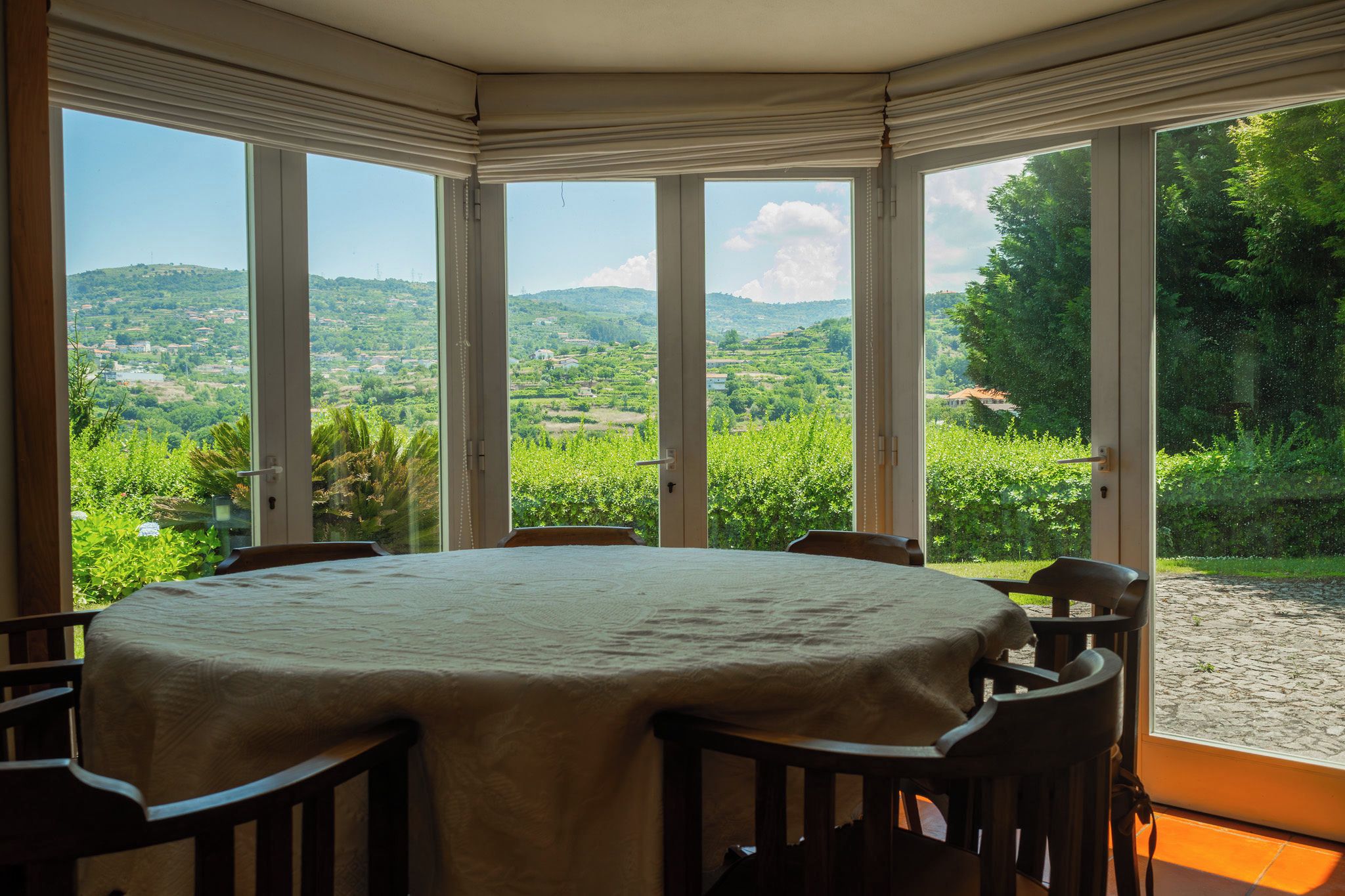 A round table with a beige tablecloth, surrounded by wooden chairs, in a room with large windows offering a view of a green landscape and hills.