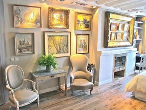 A room with a fireplace, a chair, a table with a plant, and multiple framed artworks on the wall. - A Duplex In The Sky - Paris — AC (Paris)