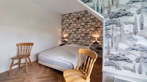 Un dormitorio con piso de espiga, una cama, dos sillas de madera, una pared con un mural temático de naturaleza y dos lámparas de noche.