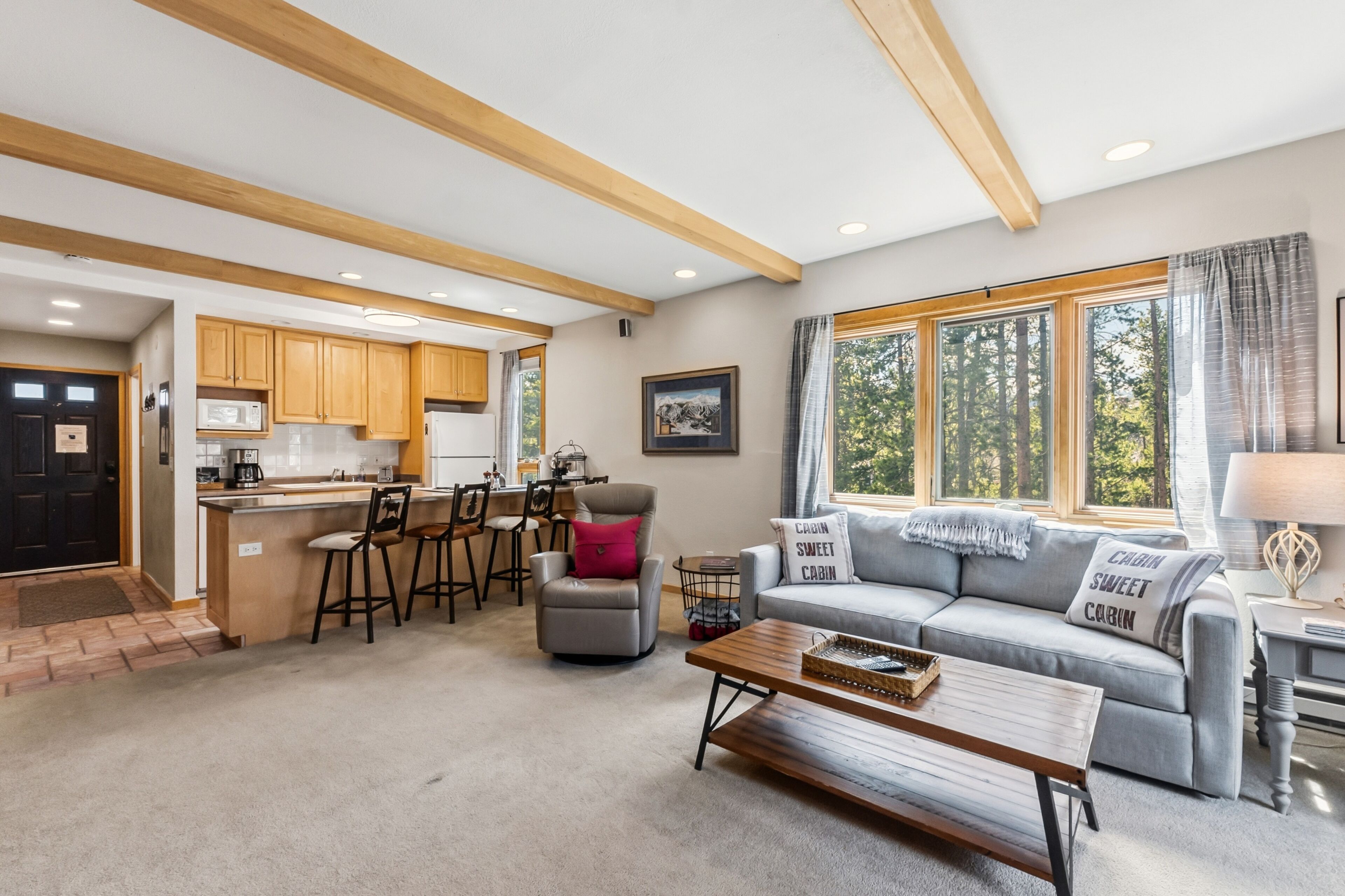 A spacious living area with a kitchen, a gray sofa with 'Cabin Sweet Cabin' pillows, and a dining area with wooden chairs and a small table.