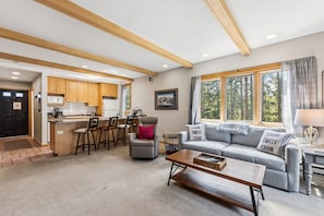 Ein geräumiger Wohnbereich mit einer Küche, einem grauen Sofa mit 'Cabin Sweet Cabin'-Kissen sowie einem Essbereich mit Holzsesseln und einem kleinen Tisch.