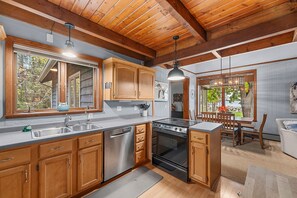 A kitchen with wooden cabinets, a stainless steel dishwasher, a black stove, and a dining area with a table and chairs. - Owls Nest: Scenic Lakefront Retreat w/ Outdoor Fun (Sturgeon Bay)