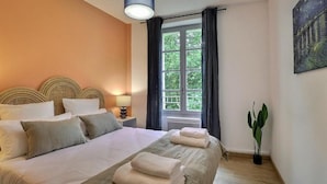 A bedroom with a large bed, a window with curtains, a bedside table with a lamp, and a wall-mounted painting. - Nuit étoilée (Arles)