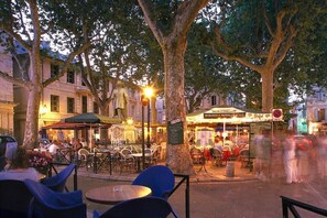 Um café ao ar livre com mesas e cadeiras, uma rua com pedestres e um edifício com uma placa.