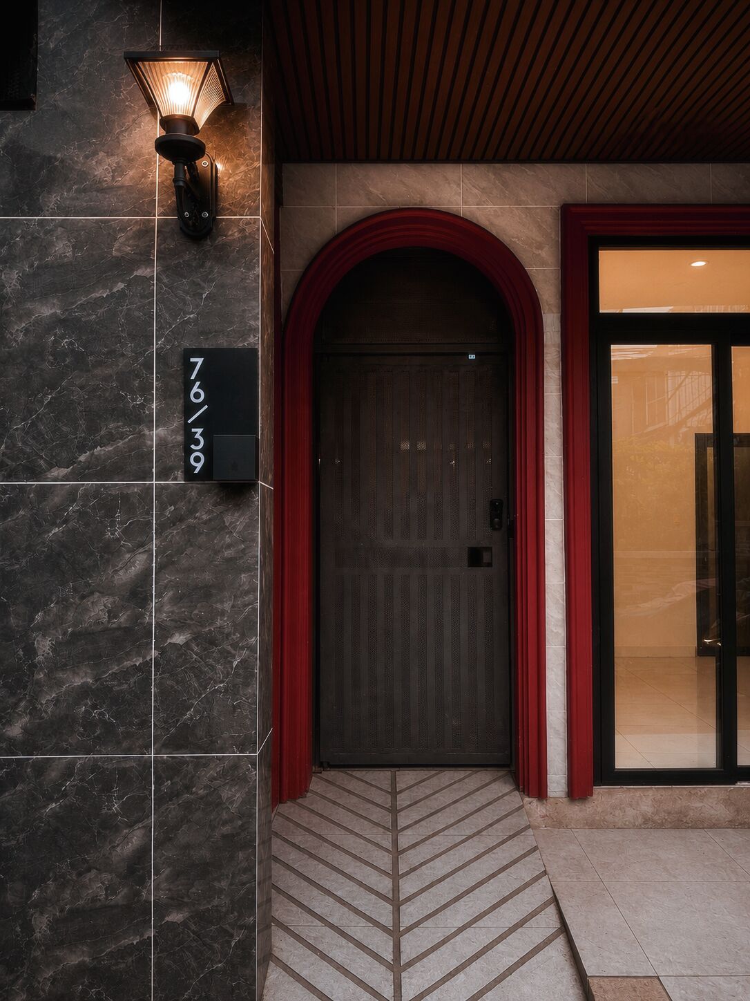 A hotel entrance with a dark marble wall, a lit wall lamp, a red-framed door, and a tiled floor.