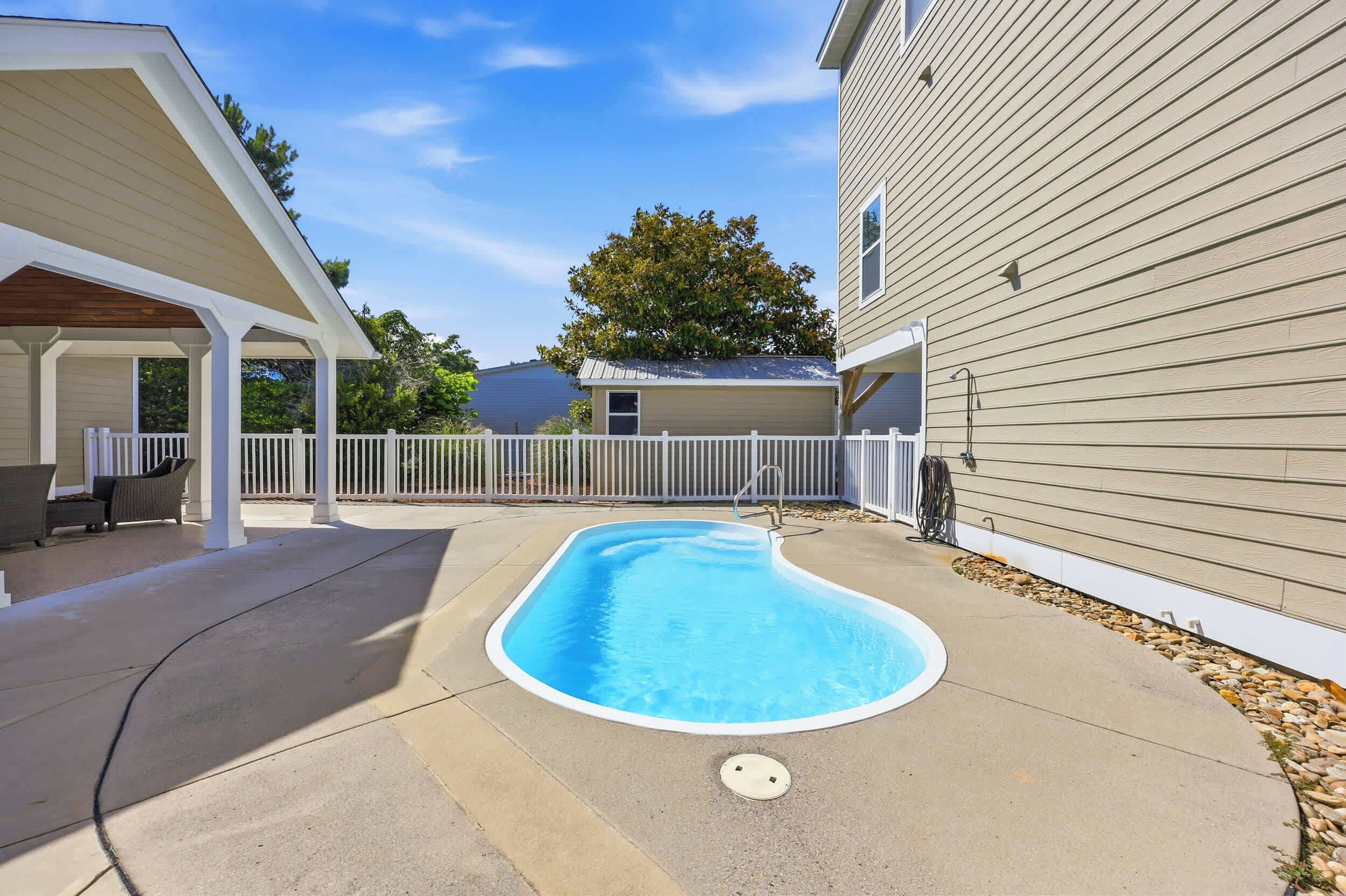 A backyard with a swimming pool, a covered patio, and a white fence.
