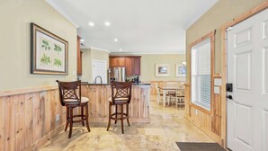 A kitchen with wooden cabinets, a central island with stools, a refrigerator, and a dining area with a table and chairs. - Sol and Sea Beach Compound w Pool and Boat House (Murrells Inlet)