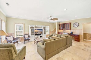 A spacious living room with a large TV, comfortable seating, a ceiling fan, and a kitchen area with wooden cabinets.
