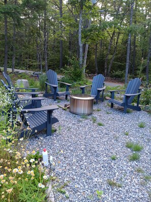 Ett utomhusområde omgivet av träd med sittplatser och Adirondack-stolar samt en eldstad.