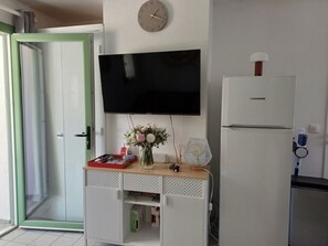 Una habitación con una televisión de pantalla plana montada en la pared, un refrigerador, una pequeña mesa de madera con un florero y una puerta con marco verde.