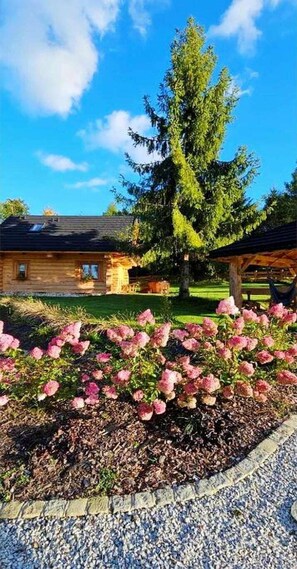Una cabaña de madera con un porche, rodeada de un jardín con flores rosas y un gran árbol.