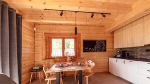 A wooden cabin interior with a dining table, chairs, a kitchen area, and a large window.