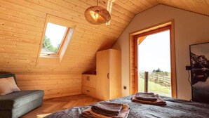A room with wooden walls, a bed with folded blankets, a sofa, a wardrobe, and a large window with a view.