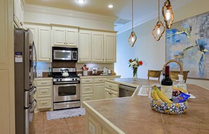 Una cocina con armarios blancos, electrodomésticos de acero inoxidable, una isla grande con fruteros y un cuadro en la pared.