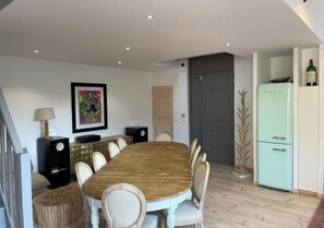 Ein Essbereich mit Holztisch und -stühlen, einem gerahmten Bild an der Wand, einem Lautsprecher, einem Kühlschrank und einer Tür.