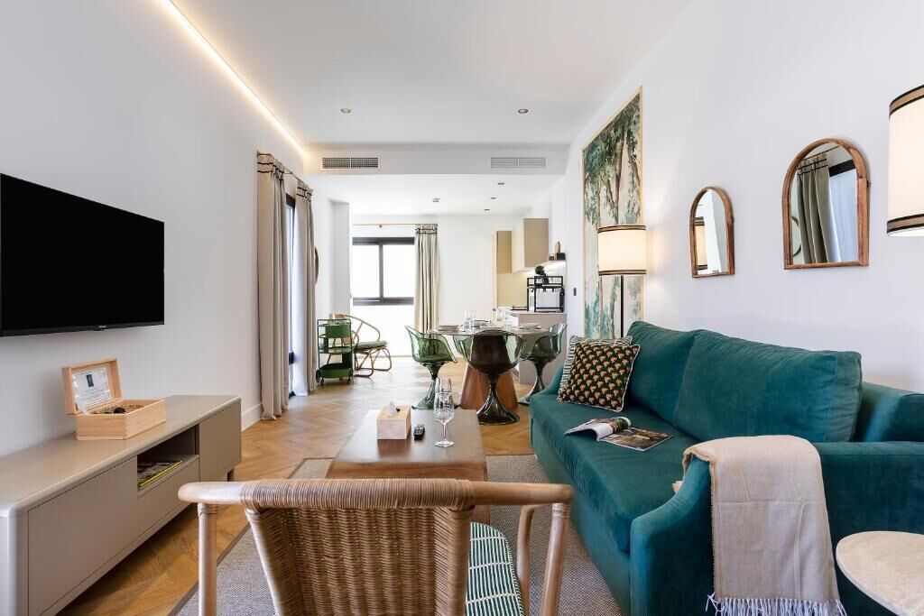 A modern living room with a flat-screen TV, a teal sofa, a dining table, and a wicker chair.