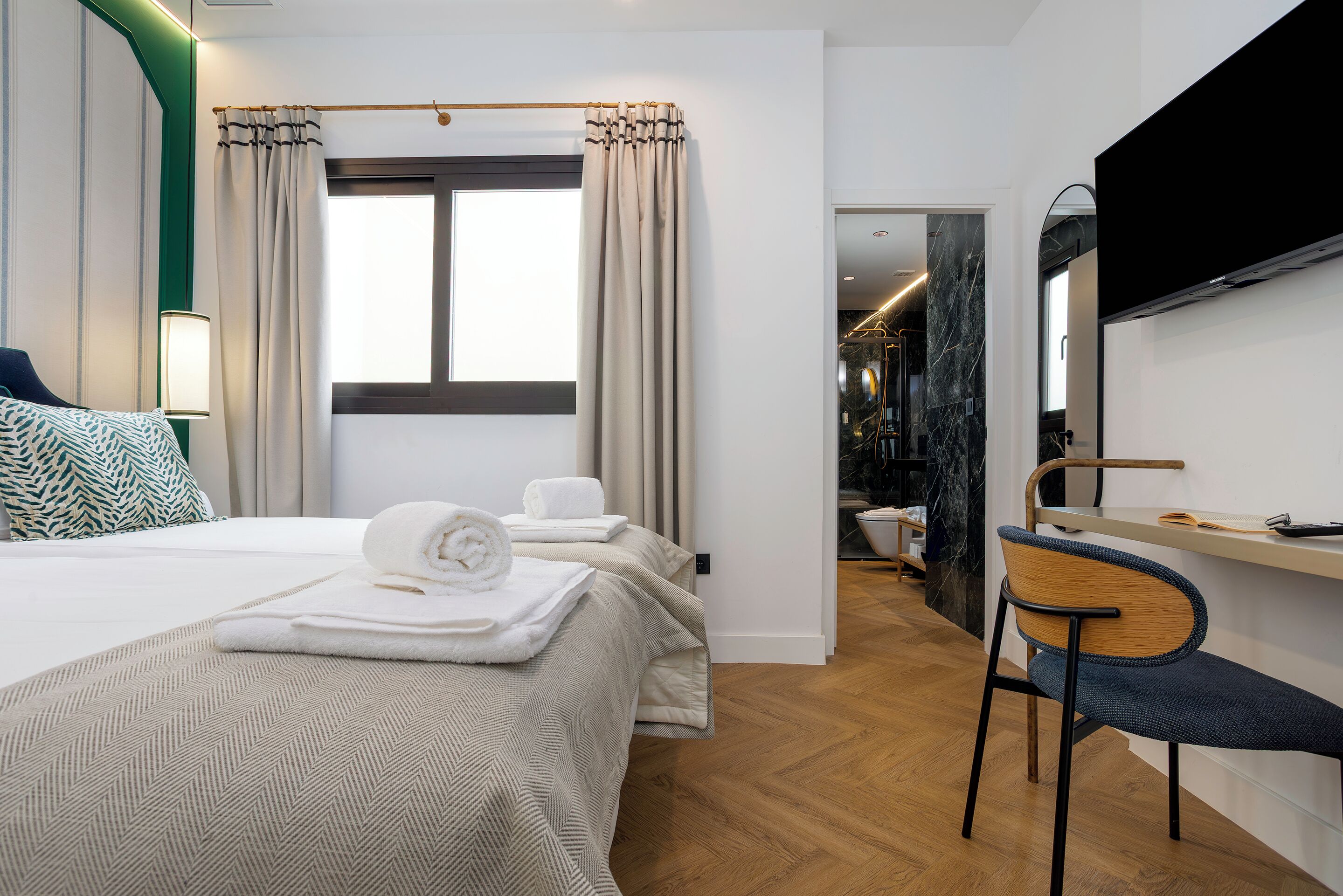 A hotel room with a bed, a chair, a desk, a television, and a bathroom visible through an open door.
