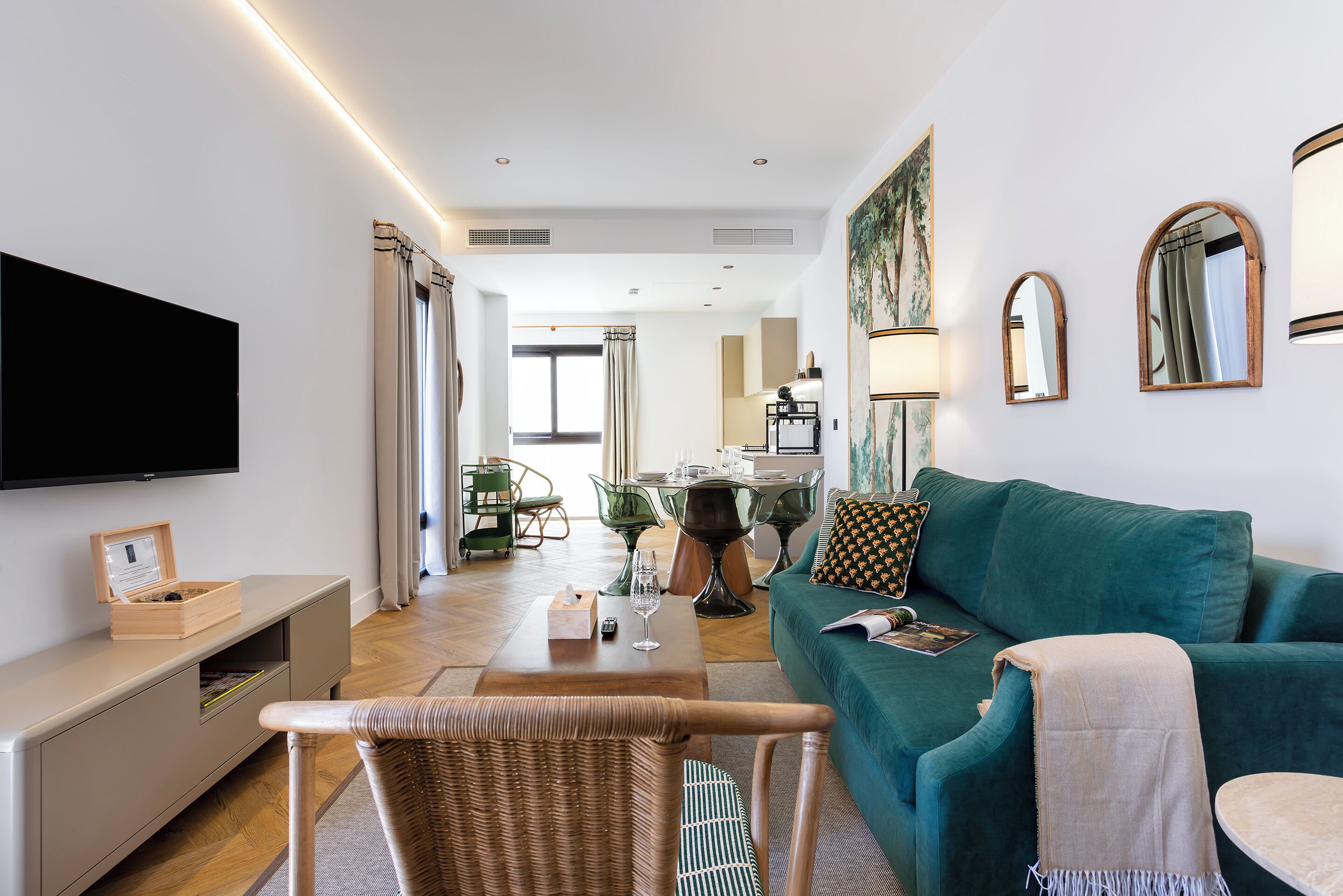 A modern living room with a teal sofa, a flat-screen TV, and a dining area with a table and chairs.