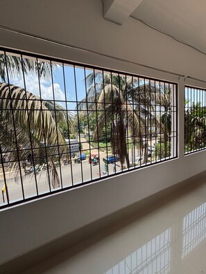 Habitación con un ventanal con barrotes de metal, techo blanco y vistas a palmeras y una calle con vehículos.