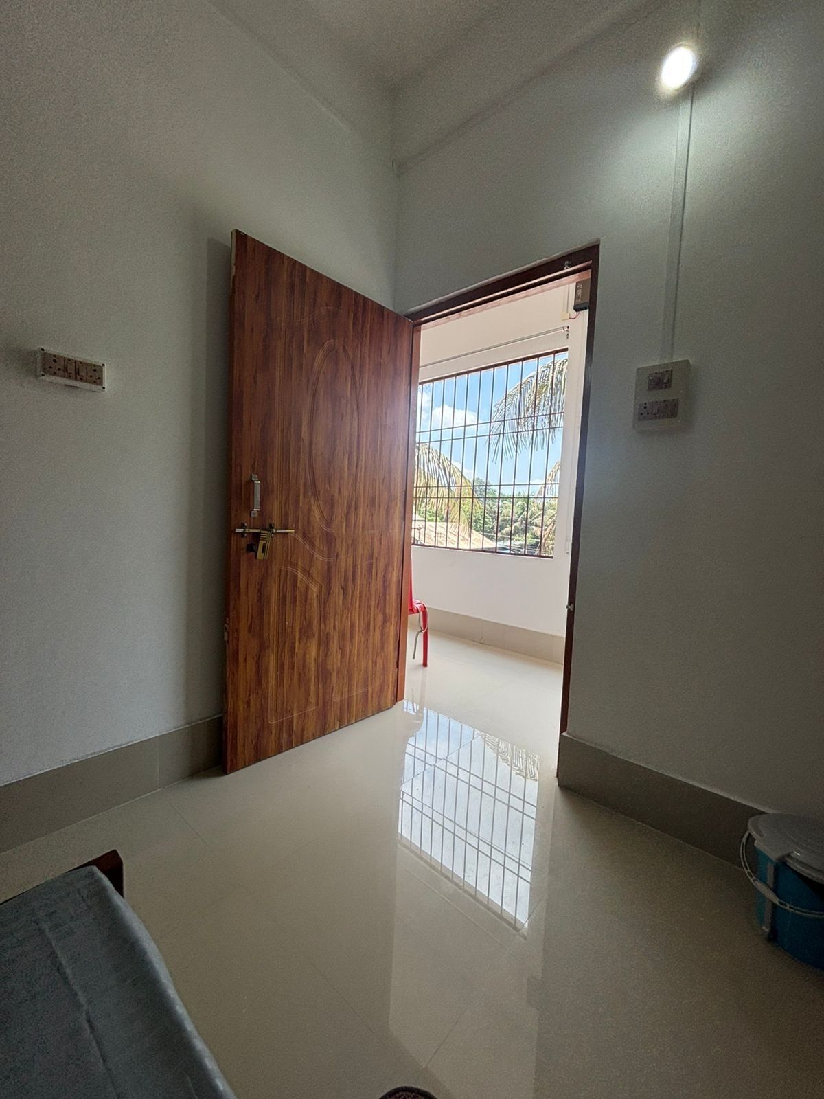 A room with a wooden door, a tiled floor, and a view through a window with a metal grid.