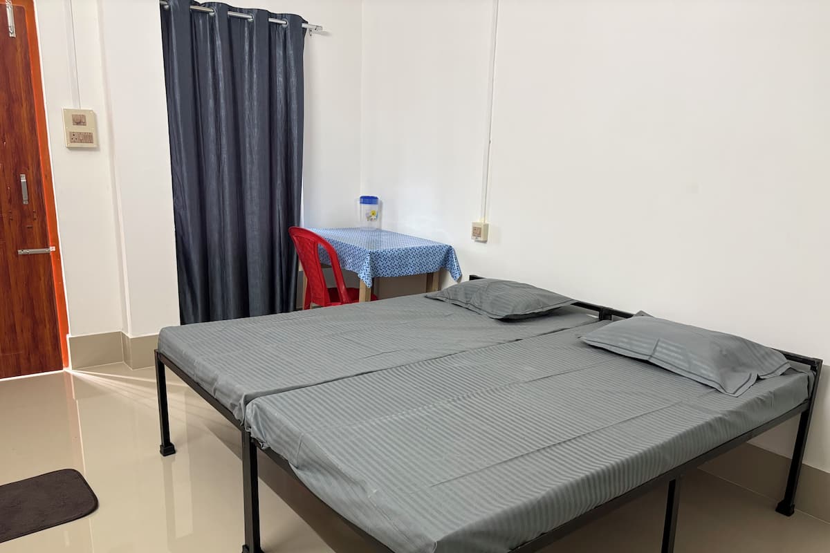 A single bed with a metal frame, a small table with a blue tablecloth, a red chair, a water bottle, and a curtain.