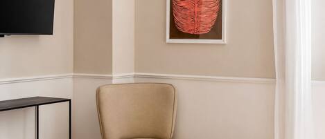 A room with a beige armchair, a small wooden side table, a framed artwork on the wall, and a black television mounted on the wall.