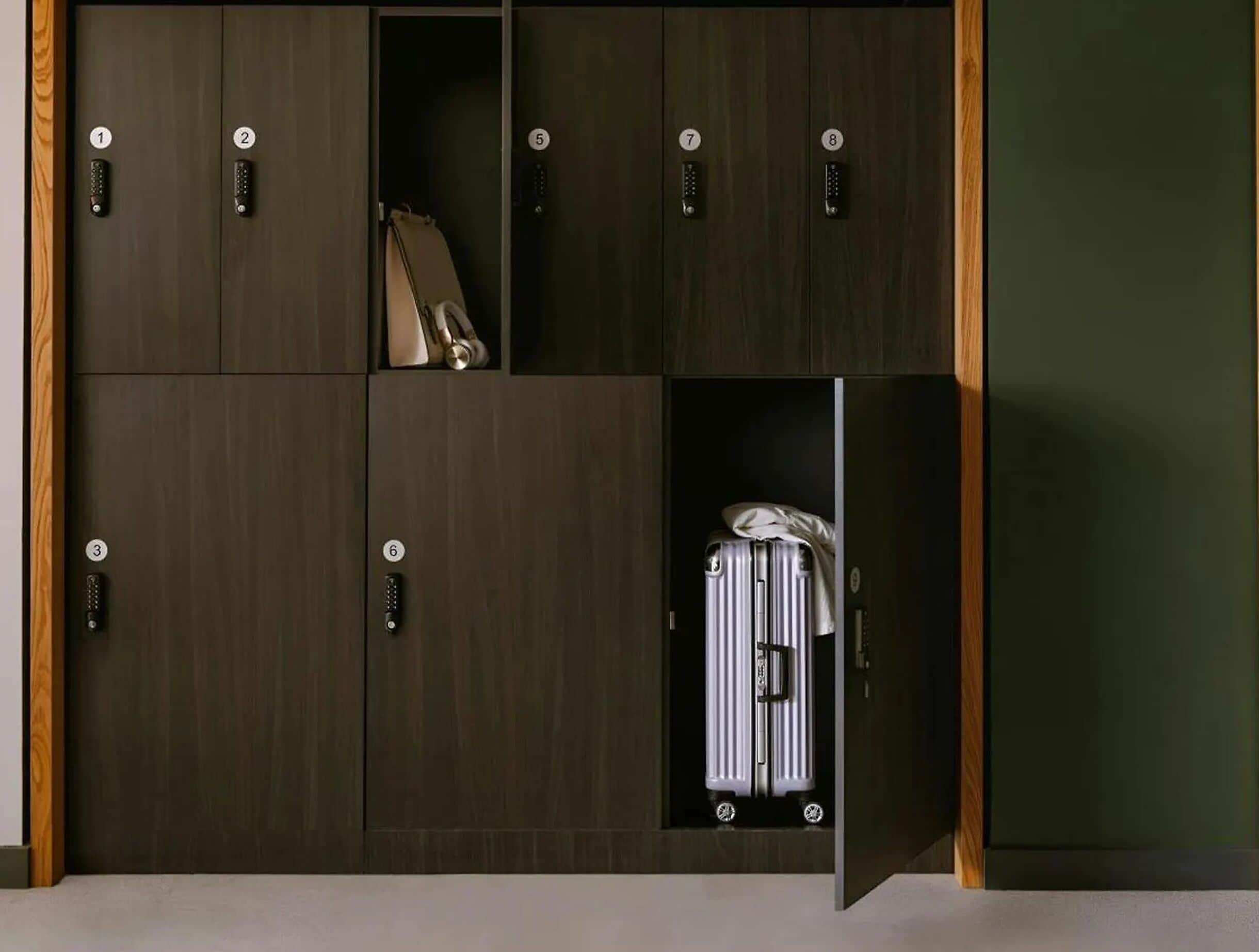 A set of dark wooden lockers with numbered handles, one of which is open revealing a suitcase.