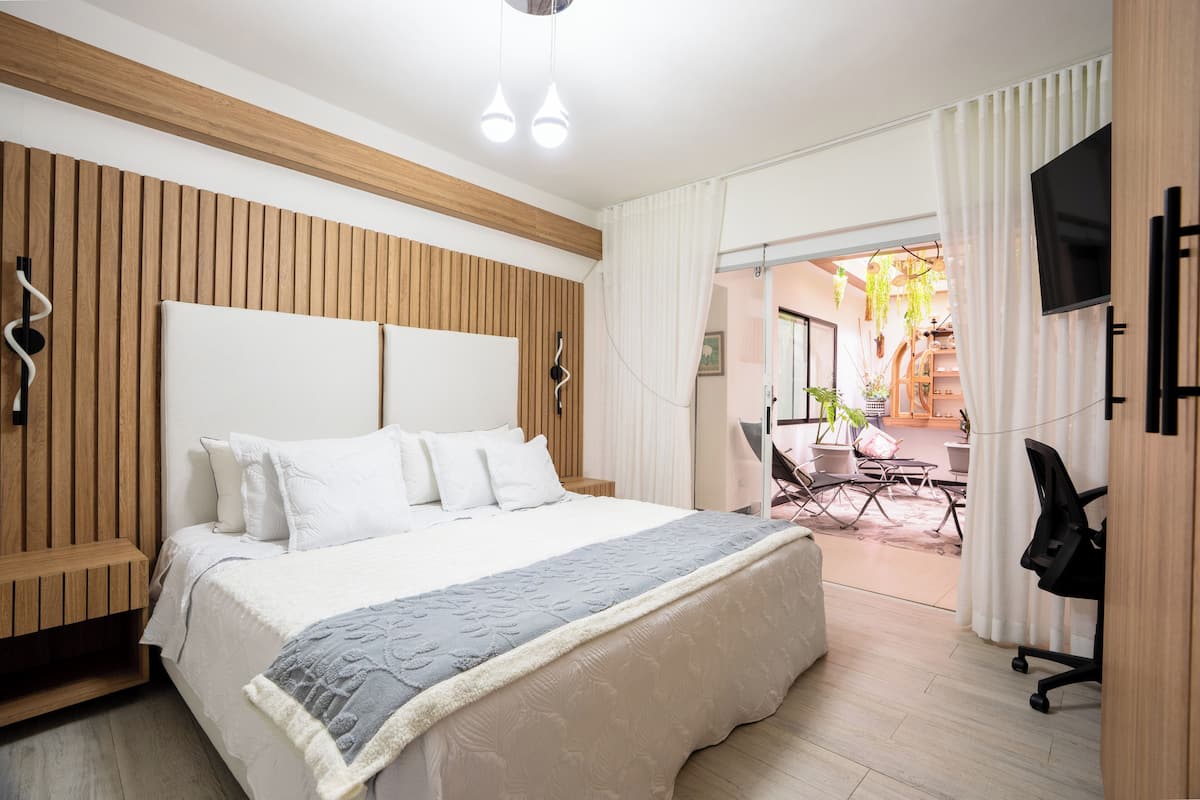 A modern hotel room with a large bed, wooden paneling, a flat-screen TV, and a balcony with outdoor seating.