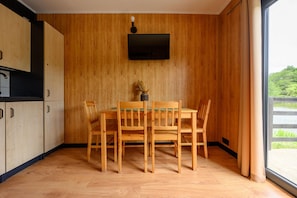 A dining area with a wooden table and chairs, a flat-screen TV mounted on the wall, and a view of greenery outside. - Single-story holiday home by the pond no. 9 (Darlowo)