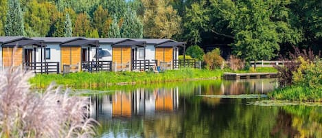 Una hilera de cabañas modernas de dos pisos con un balcón de madera al frente, rodeadas por un estanque tranquilo y abundante vegetación.