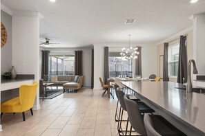 A modern kitchen with a central island, bar stools, a dining area with a sofa, and a large window. - Storey Lake 6BR Villa | Water Park + Pool Spa (Kissimmee)